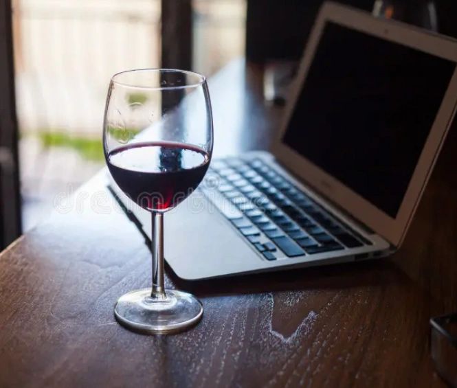 A glass of red wine beside an open laptop on a wooden table.